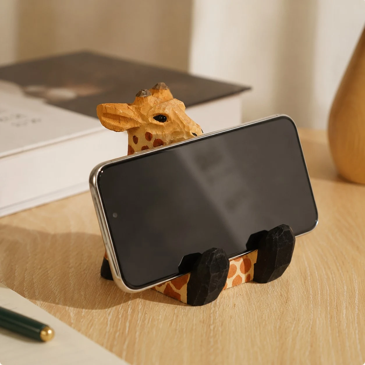Soporte de madera para teléfono con forma de jirafa que sostiene un teléfono sobre un escritorio.